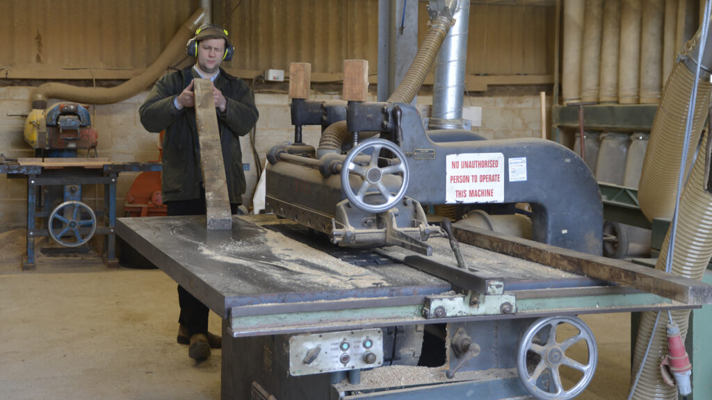 Image of a man wearing ear defenders, stood behind woodworking machinery. He is holding a long rectangular piece of raw timber, which is inspecting. The machinery is in the foreground of the image.