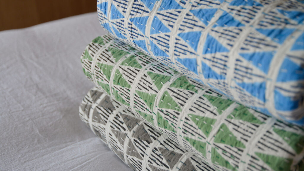 blue green and grey bedspreads with triangle print folded and stacked on a bed with white sheet.
