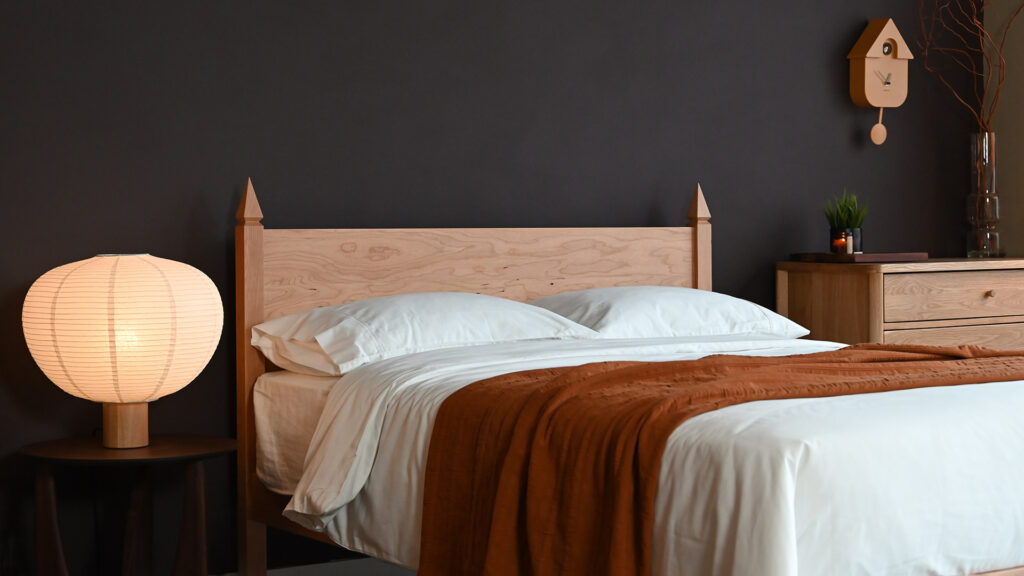 Cherry Samarkand bed base with white cotton bedding and a terracotta bedspread. Next to an oiled oak chest of drawers to the right and an oak lamp with a large paper shade to the left.