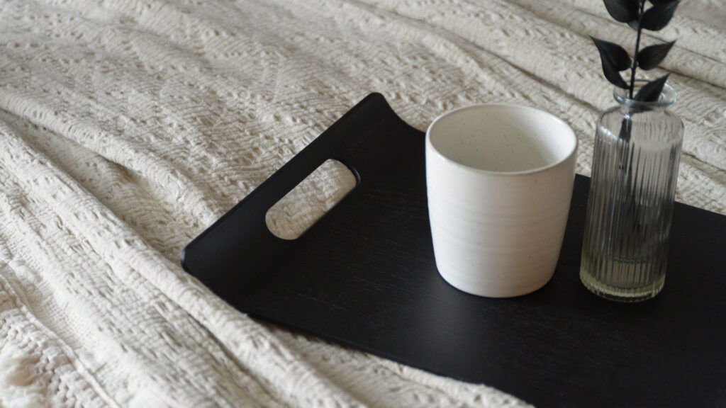 A black tray with a white mug and small glass vase photographed on a white textured bedspread.