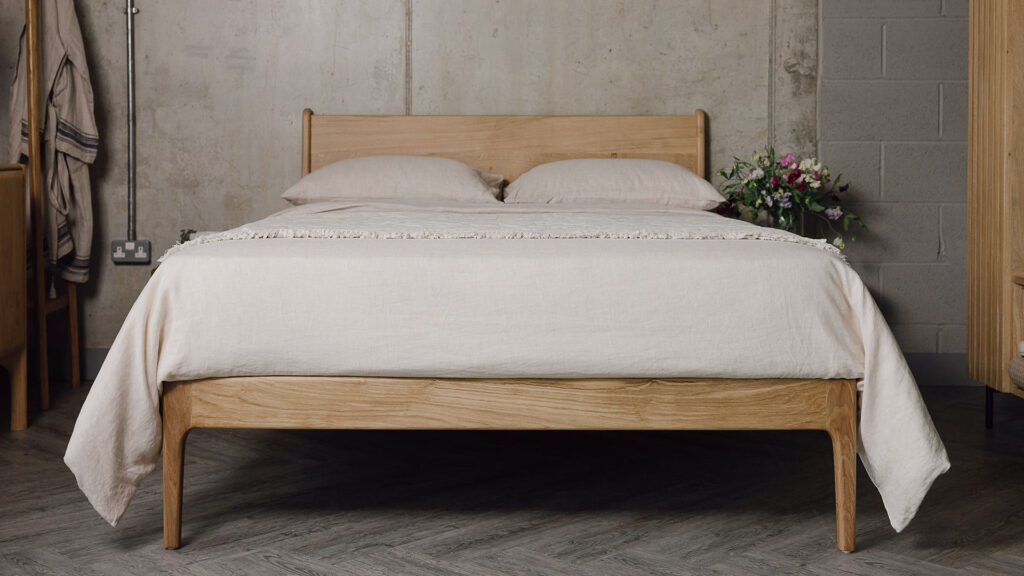 A shot of the English Oak Camden, showing its headboard and focusing on its foot-rail primarily. The bed base is dressed in the organic linen bedding and the floral textured throw in the ecru colour-ways.