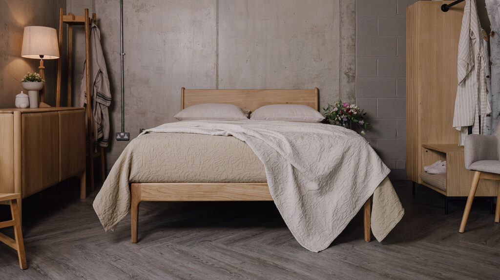 Zoomed-out shot of the English oak Camden, showing its headboard and foot-rail. It is dressed with the floral textured bedspreads in the colours ecru and sand. On one side of the bed sits the Ethnicraft oak Pi sideboard and the Tower oak coat-stand, and on the other side of the bed sits the Modern Oak Open Wardrobe.