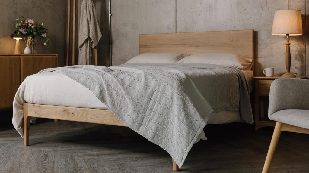A low angled shot of the English oak Chiswick, dressed in the organic linen bedding in ecru. The bedding is mostly covered up by a embroidered striped bedspread in the colour silver-grey, which also overhangs the bottom right-hand corner of the bed base.