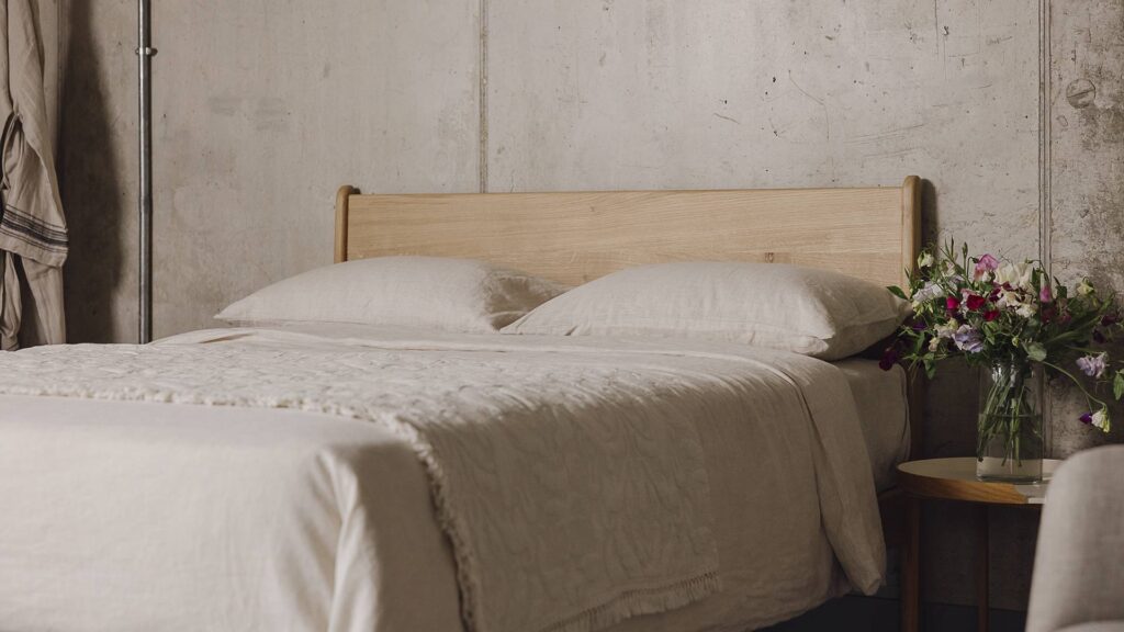 Zoomed out shot of the English oak Camden's headboard panel. The bed is dressed in the organic linen bedding and the floral textured throw in the ecru colour-ways. There is a round-topped table next to the bed with a clear vase full of colourful flowers.