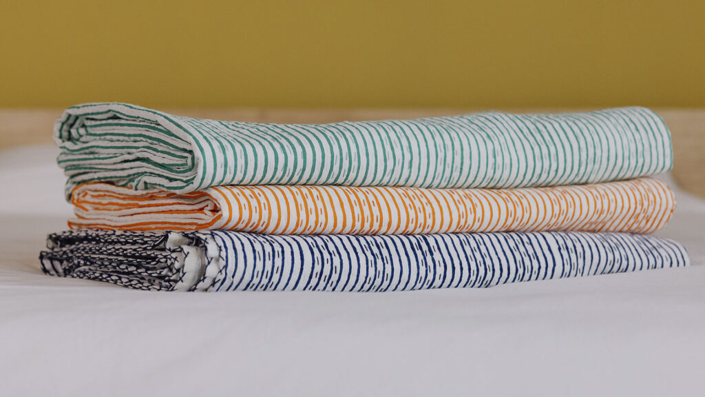 3 striped kantha bedspreads neatly folded and stacked on a white sheet. Top to bottom - green, orange and dark blue.