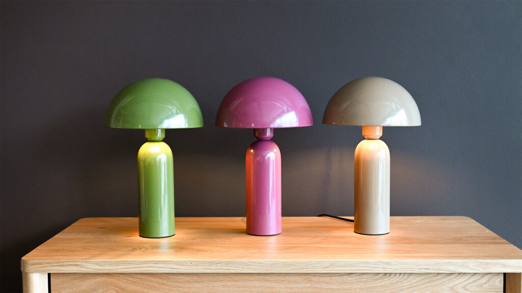 Three retro style lamps displayed atop an oak chest of drawer to show the range of colours available. The colours are green, mauve and mocha brown.