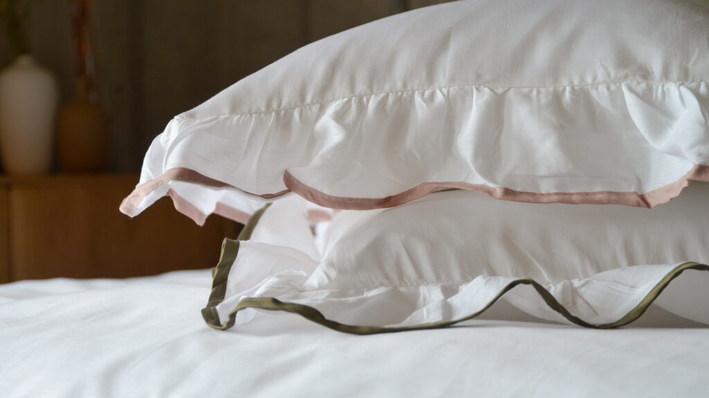 A stack of two pure white pillowcases with ruffles edges. Once with a dusty pink edge, the other olive green.