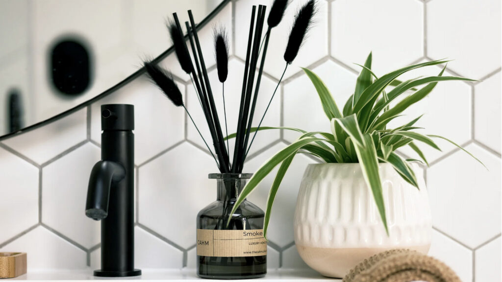 Smoked cedar diffuser in dark glass with black reeds and bunny tails displayed on bathroom side with black tap and a houseplant.