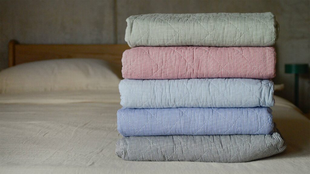 stack of five folded quilted striped throws. Green, red, pale blue, dark blue and dark grey.
