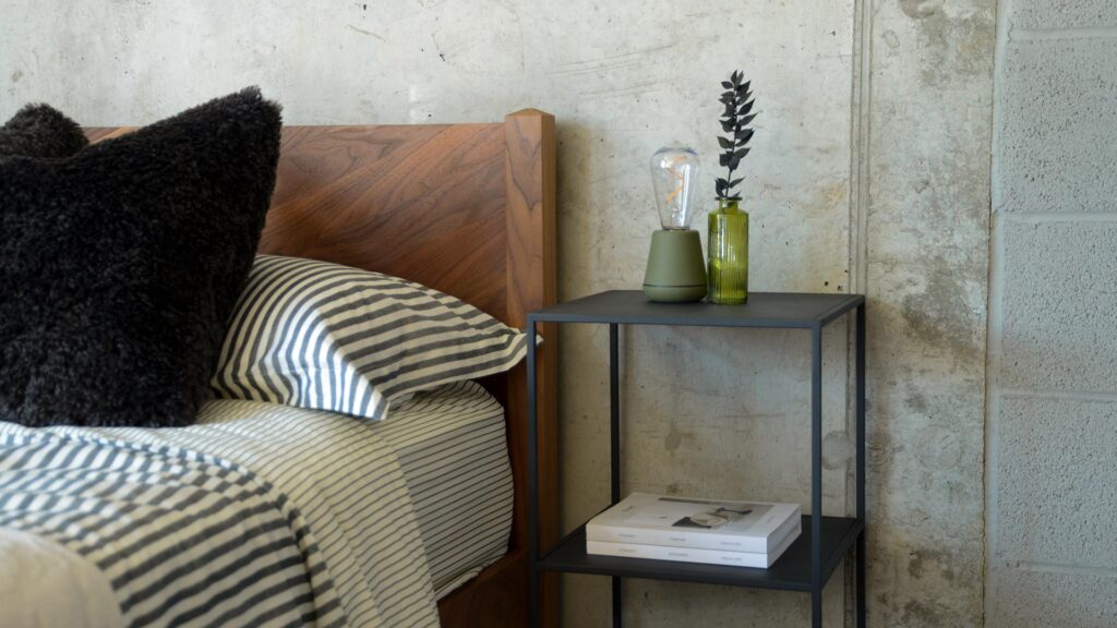 Image showing Fera tall side table in powder coated metal in a dark grey charcoal colour next to a walnut bed base.