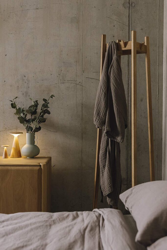 Portrait image showing the Tower Coat stand with a clay coloured organic cotton robe hanging from it. To the left, you can also see part of a solid oak unit from Ethnicraft, with some small accessories - including a lamp and vase - sat atop it. To the bottom of the image, you can see a bed base, but this is not the main focus of the image.