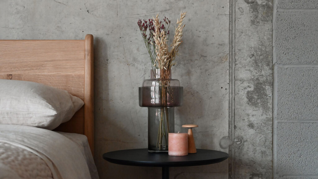 Two tone glass vase with removable top and narrow opening on a black bedside table with a small lamp and candle. Small bouquet in vase.