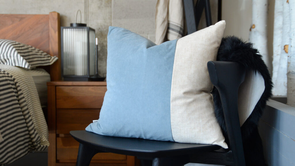 A pale blue Velvet Block Cushion sat on a black chair with a black sheepskin over the back of it. The chair is sat in a bedroom setting with a walnut bedside table and walnut bed visible in the background. The cushion is the focal point of the image and is made from velvet and linen.