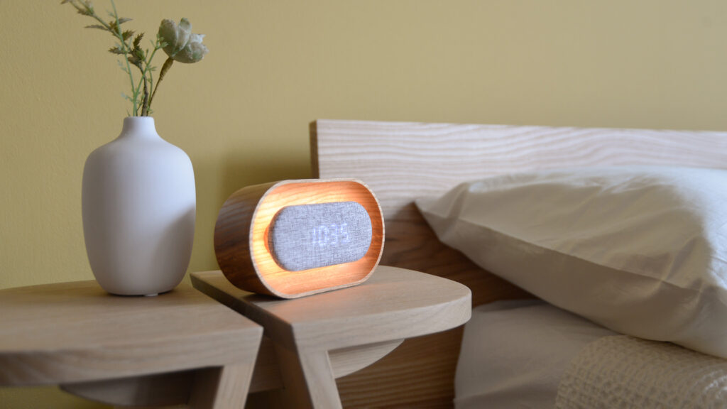 side view of lit up oak alarm clock on bedside table next to a white vase.