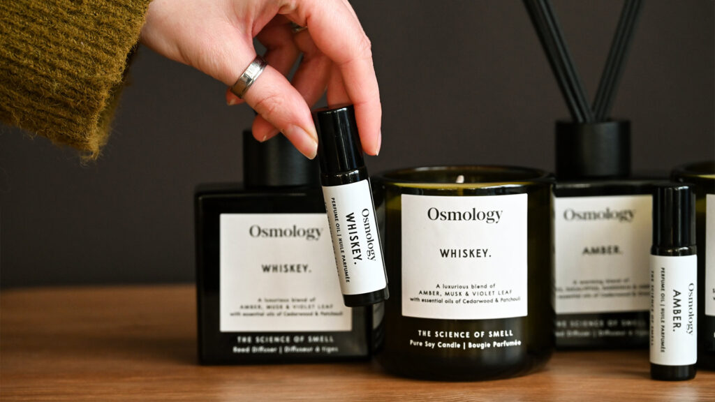 A close up image of a hand holding up the Aery Osmology roll-on perfume oil in the 'Whiskey' scent. In the background, the Aery diffusers, roll-on perfume oils and candles are visible in the Whiskey and Amber scents.
