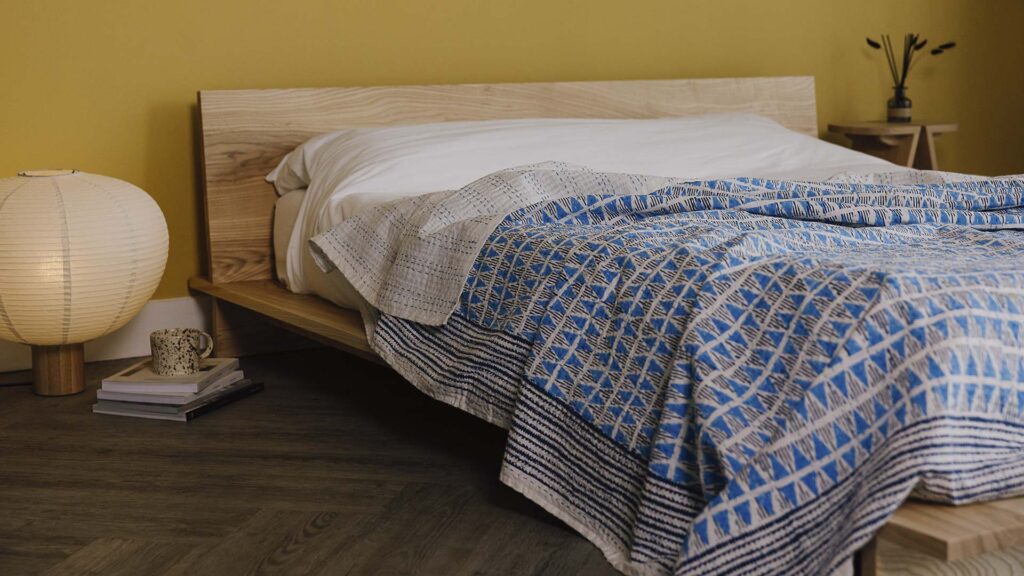 ash Oregon bed with white linen and a blue block print bedspread. Bedroom scene also features a bedside table, large lamb, stack of books and a mug.