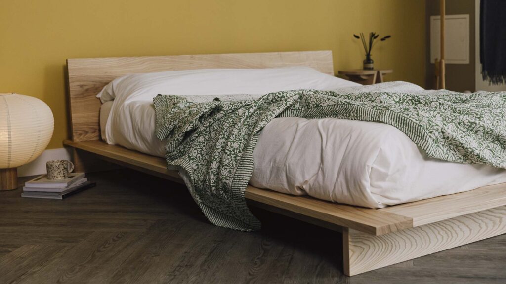 green floral bedspread messily draped over white bedding on an ash bed frame.