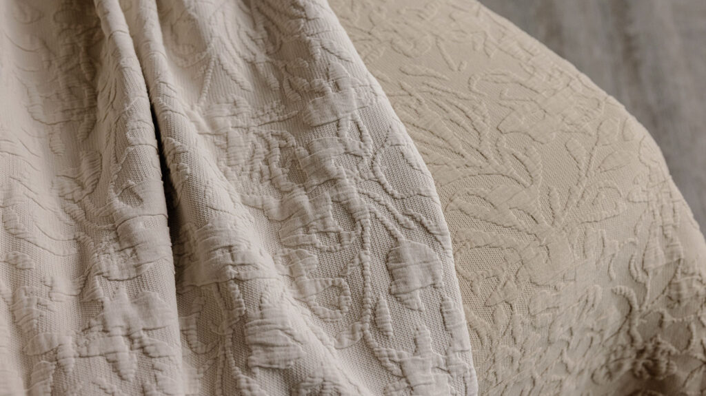 A close up image of an ecru coloured floral textured bedspread laid on top of a sand coloured floral textured bedspread.