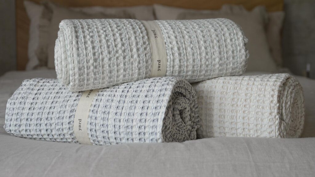 Close up of three rolled up waffle throws. One ivory, one ivory with navy detail, one ivory with green detail.