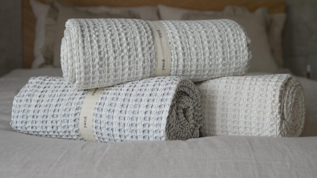 Close up of three rolled up waffle throws. One ivory, one ivory with navy detail, one ivory with green detail.