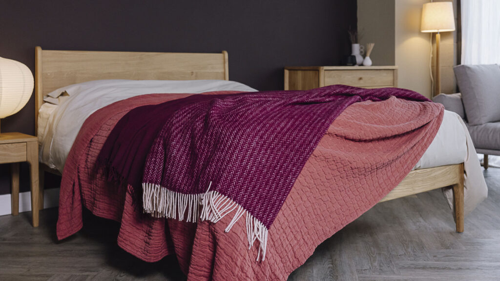 Bedroom scene with oak bed frame and furniture. Bed dressed in raspberry bedspread, with a deep burgundy plain throw, and herringbone patterned throw with white tassels over the top.