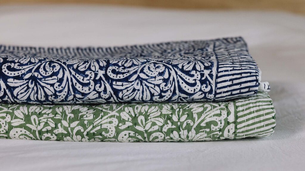 Stack of 2 floral kantha bedspreads folded neatly. Botton green, top blue.