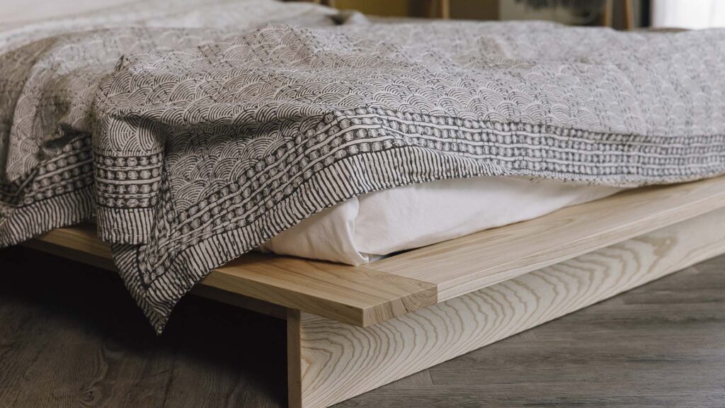 bottom corner of a platform bed frame in solid ash. A grey shell print bedspread draped over the top.