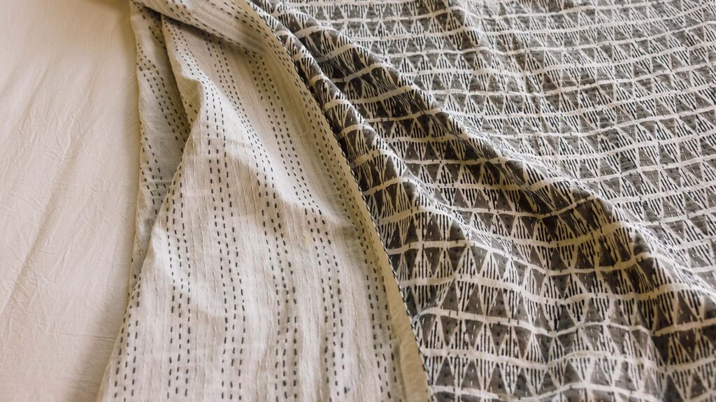 close up of a grey triangle pattern block print bedspread, top folded over to show white material with grey stitch detailing on the reverse.