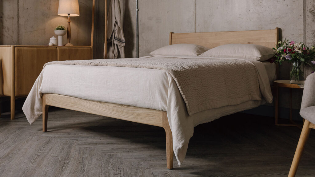 3/4 view of a solid English oak bed with ecru bed linen. A quilted velvet bedspread neatly draped over the top.