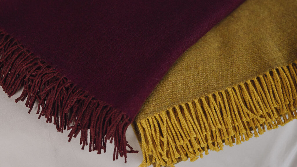 2 Rich merino throws with tassels neatly folded on bed. One mustard yellow, one deep wine.