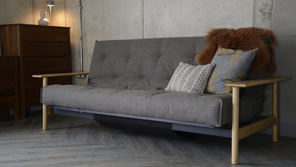 Mid-century sofa bed with oak arms. The upholstery is in the colour 'Kenya Taupe' which is a mid grey-brown colour. There are also cushions and a sheepskin decorating the seat of the sofa bed.