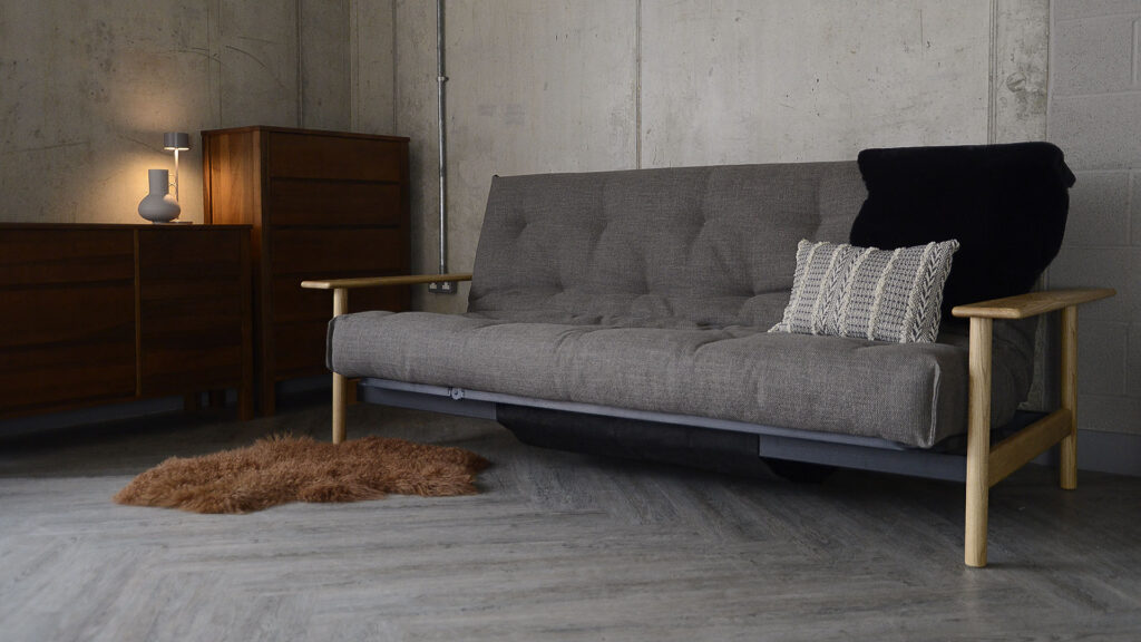 Wide angle shot of the mid-century sofa bed with a sheepskin rug on the floor and a cushion and faux sheepskin on the sofa itself. The sofa bed is sat against a concrete wall and has some dark walnut furniture to the right.