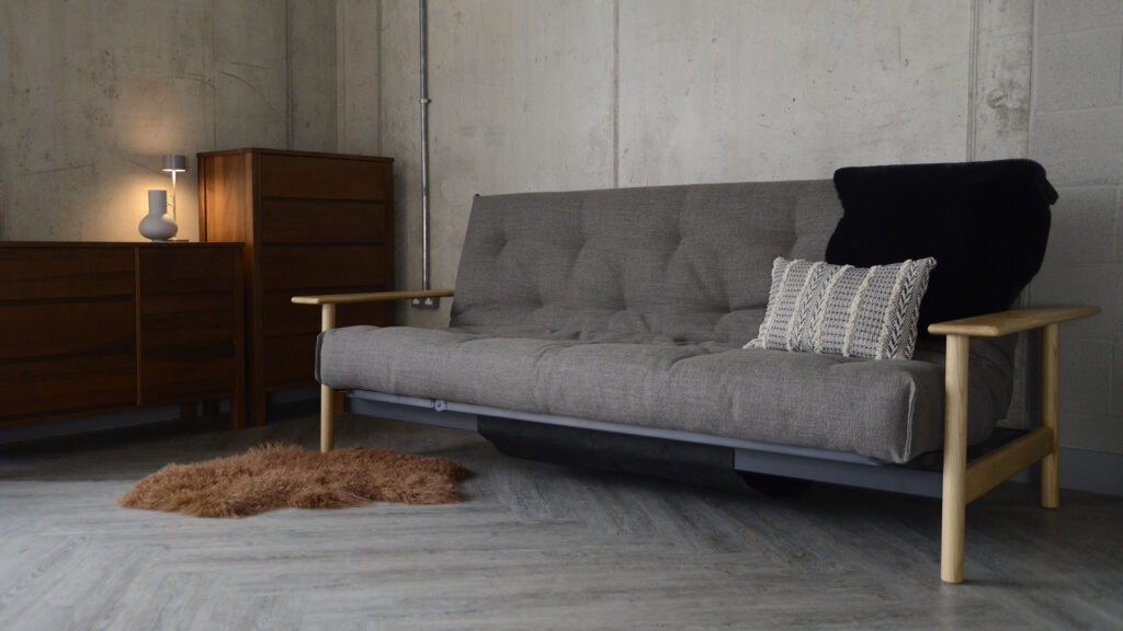 Wide angle shot of the mid-century sofa bed with a sheepskin rug on the floor and a cushion and faux sheepskin on the sofa itself. The sofa bed is sat against a concrete wall and has some dark walnut furniture to the right.