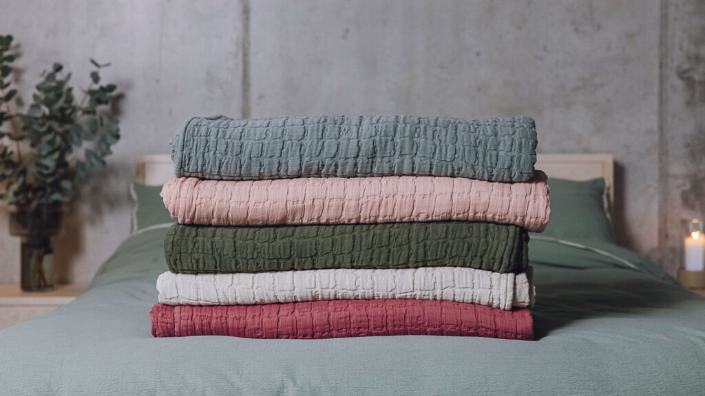 Stack of 5 textured bedspreads neatly folded on a bed. From bottom to top, raspberry, light grey, dark green, soft pink and eucalyptus.