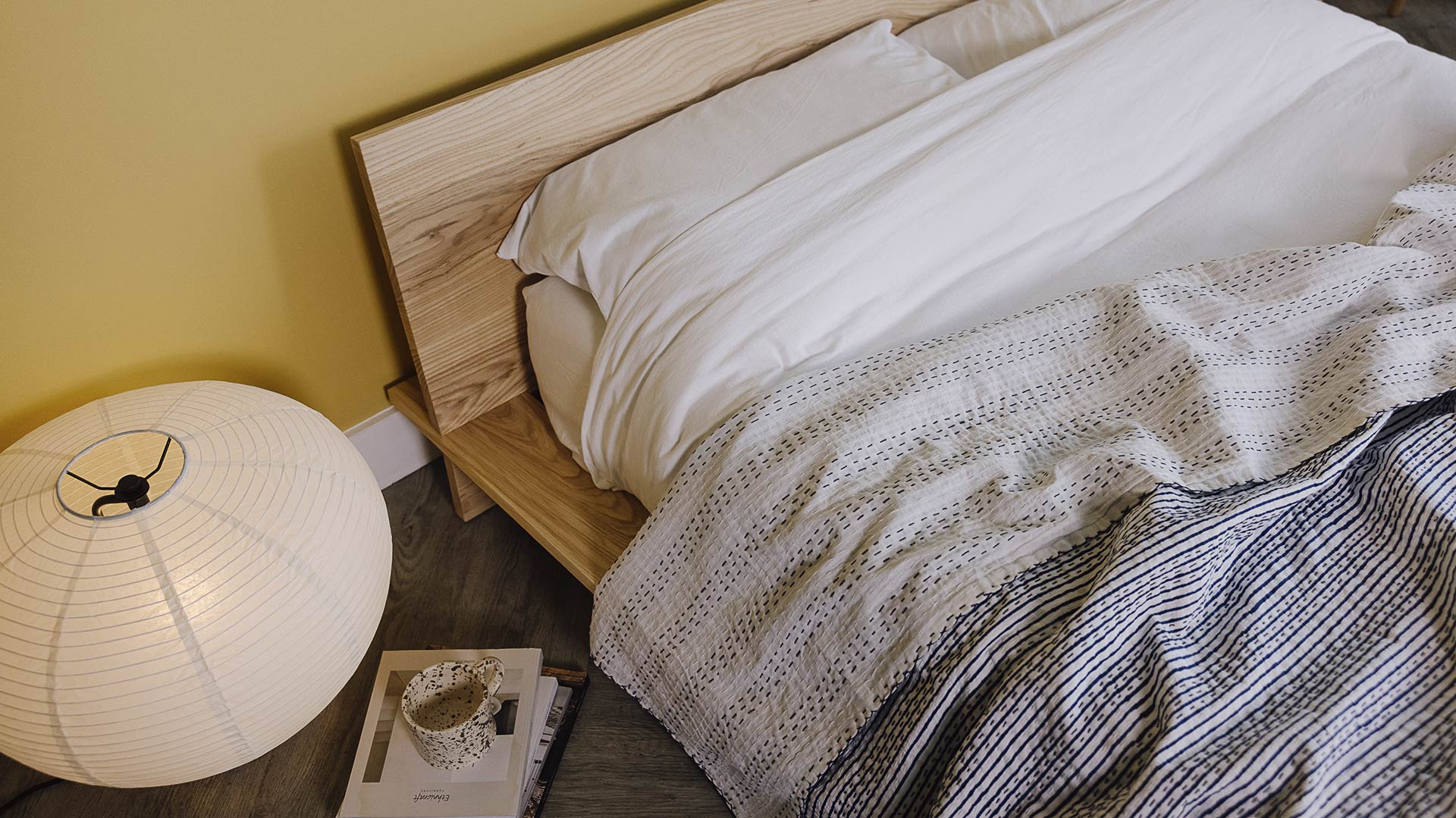 Image looking down on a modern loft bed in ash. Dressed in white linen with a blue and white striped bedspread draped over the top. A large Japanese style lamp, stack of books and mug on the floor to the side.