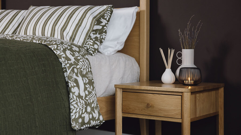 Oak bedside table with vase, diffuser and candle holder next to an oak bed with green patterned linen. Close up.