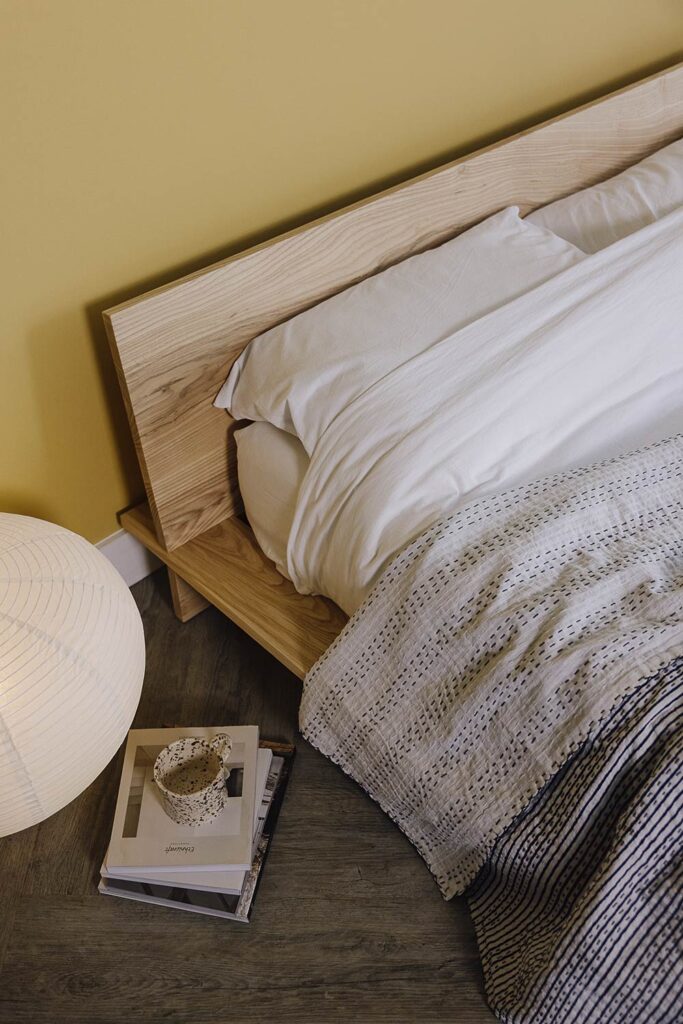 Image looking down on an ash platform bed with white linen and a blue and white bedspread. Lamp and a stack of books on the floor to the side.