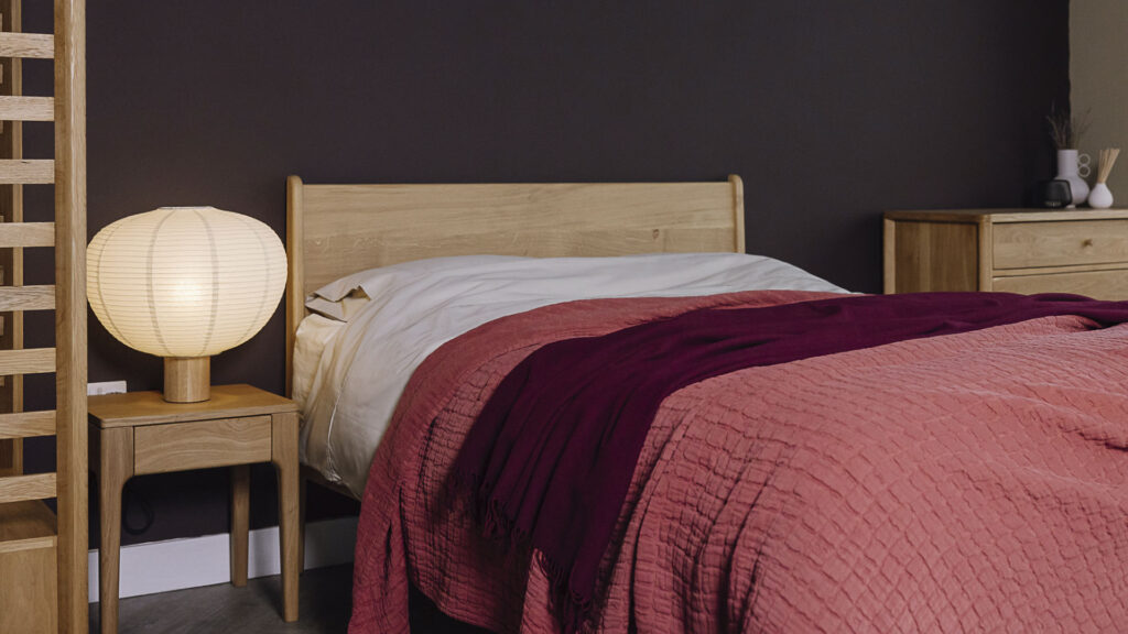 Bed scene with oak furniture. Bed dressed with off neutral linen, topped with a raspberry textured bedspread and a deep burgundy throw.