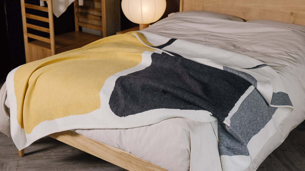 Reverse of charcoal and mustard blanket with block colours draped over bed.