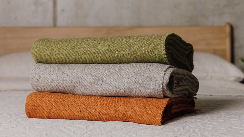 Stack of three lambswool merino throws on a bed. Top to bottom - Green, griege, burnt orange.