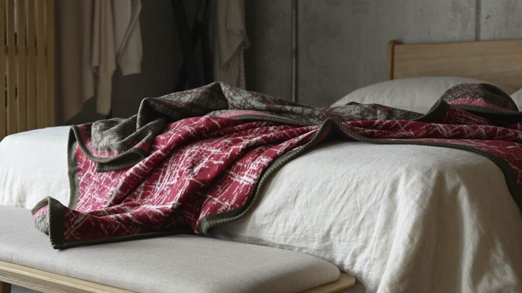 Red throw with white crosshatch pattern and Olive border messily throw over a bed with stone linen.