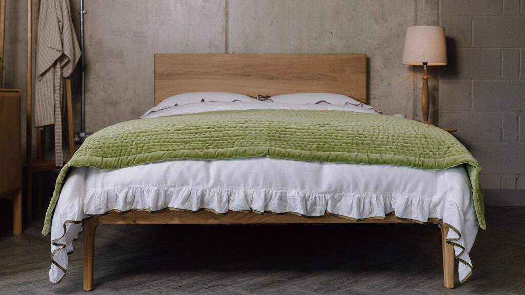 A low, forward facing shot of the English oak Chiswick, dressed in white bedding with a ruffled border that has green detailing. Over the top of the bedding is a green stitched velvet throw.