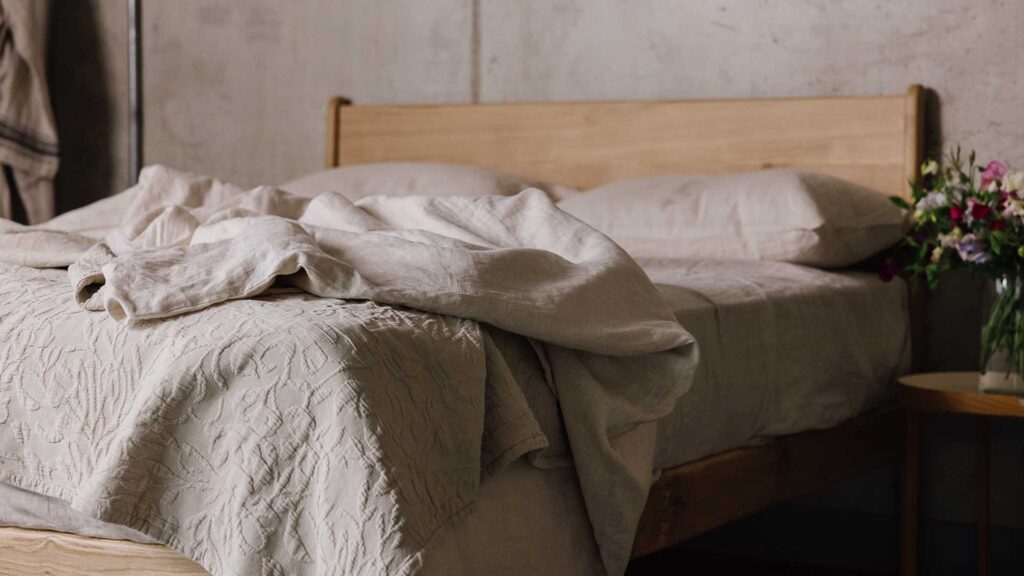 close up of bottom corner of English oak bed with messily folded back ecru bedding and textured throw.