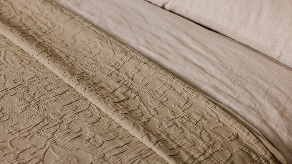 Close up shot of the sand coloured floral textured bedspread overlapping the organic linen bedding in ecru.