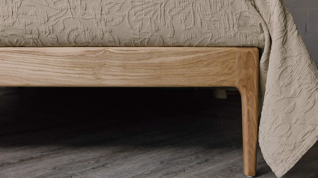 Close up of the English oak Camden's foot-rail. The bed is dressed in the sand-coloured floral textured bedspread.