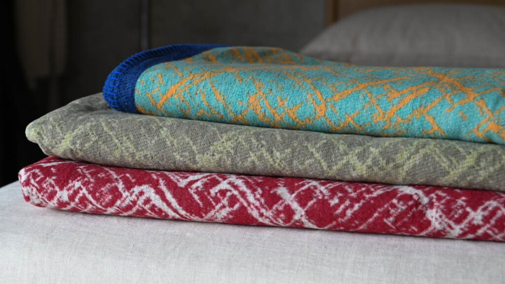 three neatly folded and stacked throw. Bottom red with white crosshatch pattern, middle shades of green, Top teal with yellow.