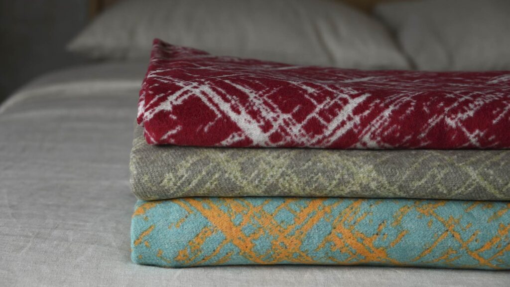 three neatly folded and stacked throw. Top red with white crosshatch pattern, middle shades of green, Bottom teal with yellow.