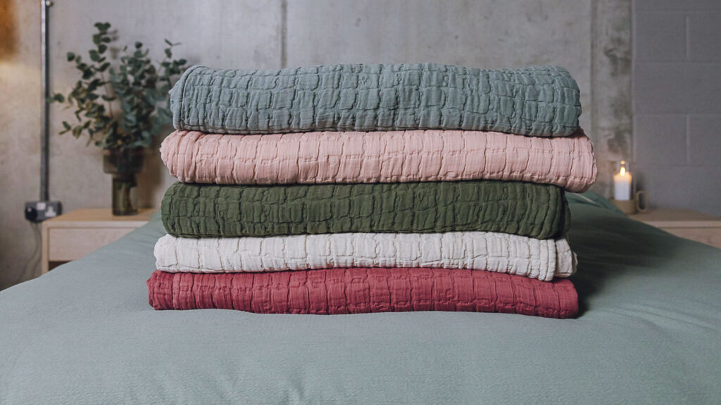Stack of 5 textured bedspreads neatly folded on a bed. From bottom to top, raspberry, light grey, dark green, soft pink and eucalyptus.