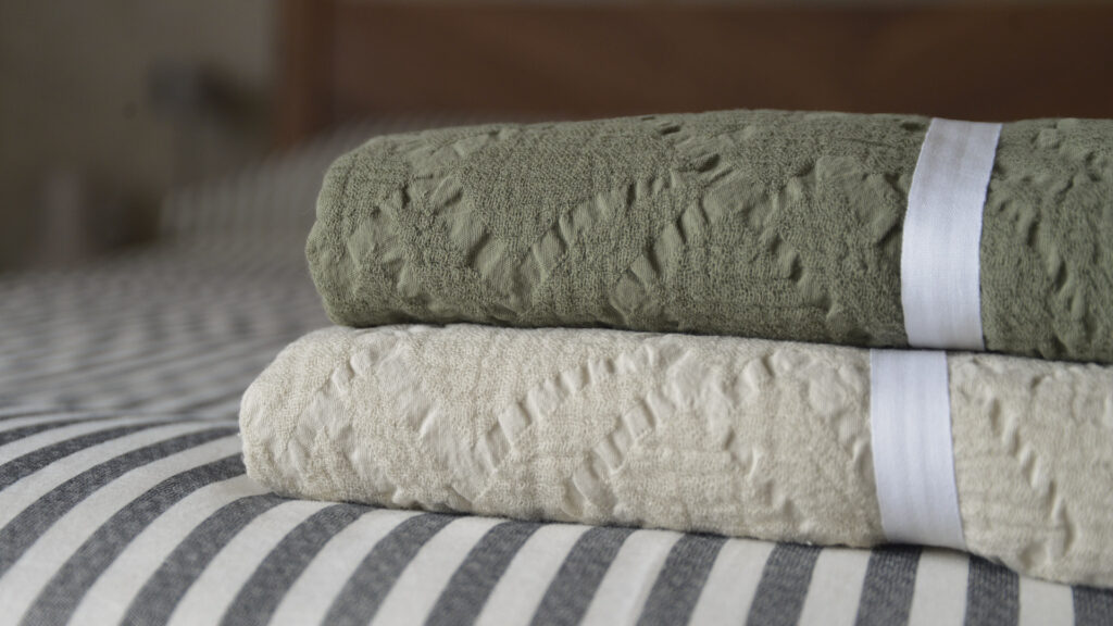 Stack of folded textured cotton throws, showing olive green on top of light ecru, laid on top of black and white stripe bed linen.