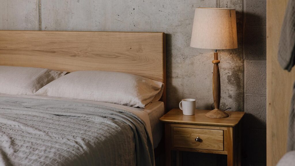 An image of part of the English oak Chiswick, showing its headboard panel. The bed is dressed in the organic linen ecru bedding and a striped embroidered bedspread in silver-grey. Next to the bed, there is a Classic Oak Nightstand. The table has a hardwood lamp with a beige lampshade and a beige mug sat on top of it.